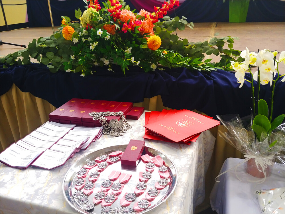 Na pierwszym planie medale Prezydenta RP za "Długoletnie pożycie mzałżeńskie", listy gratulacyjne od Gminy Raków, w tle duży bukiet kwiatów oraz udekorowana scena. 