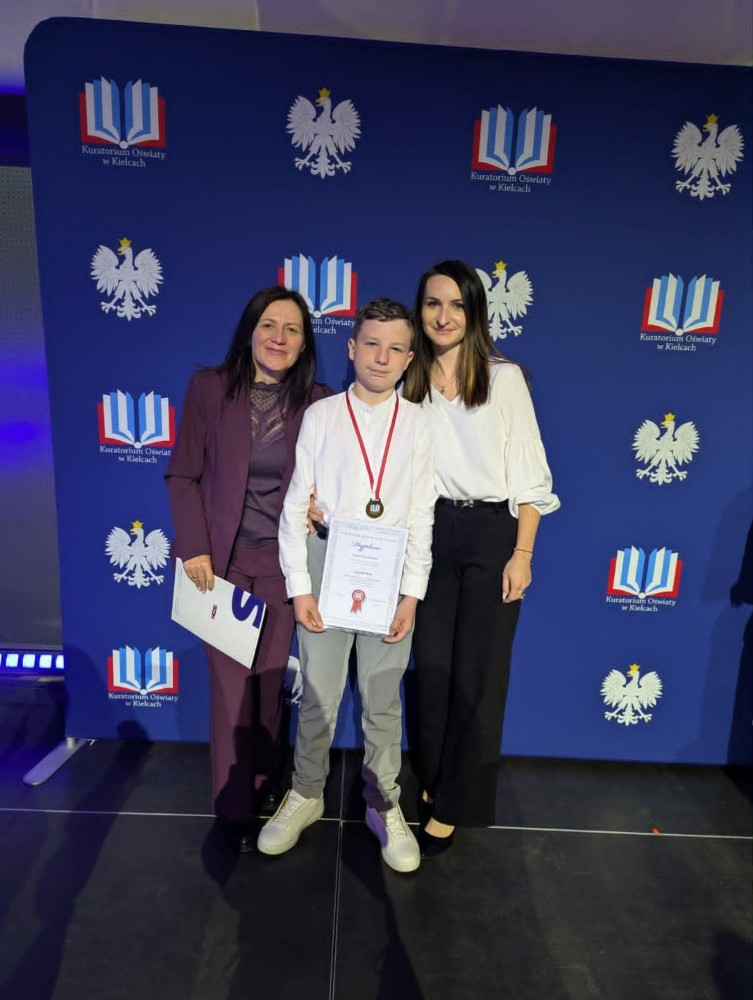 Zdjęcie przedstawia chłopca z medalem i dyplomem stojącego pomiędzy dwiema kobietami podczas uroczystości szkolnej. W tle widoczna jest ścianka z logo Kuratorium Oświaty w Kielcach oraz godłem Polski.
